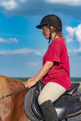 A young beautiful girl in a jockey cap and a red T-shirt rides a horse.