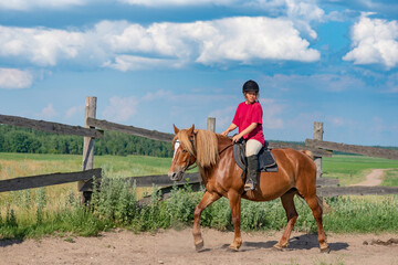 A young beautiful girl in a jockey cap and a red T-shirt rides a horse.