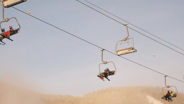 Skiers on chair lift move, climb up on sunny winter morning, backdrop of blue sky, pine forest in snow. Bottom view. People are resting at ski resort. Cableway lifts skiers in golden sunlight haze