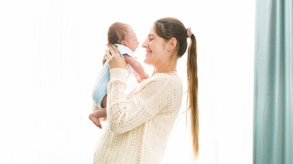 Happy smiling woman with little 1 months old baby boy hugging and embracing at big windw in bedroom. Concept of family happiness and loving parents with little children