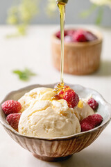 Homemade caramel vanilla ice cream with flowing syrup, frozen raspberries in ceramic bowl standing on white marble table with flowers behind.
