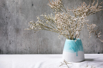 Dry white flowers branch in white ceramic vase on white linen tablecloth with gray wall behind. Copy space.