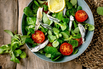 Green field salad with pickled anchovies or sardines fillet, and cherry tomatoes, served in blue bowl with lemon and olive oil on straw napkin over old wooden background. Flat lay, space