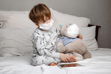 Sick five-year-old boy in a medical protective mask plays with his favorite toy while lying on a bed at home . Concept of a quarantine and epidemic strain of coronavirus.