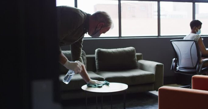 Senior caucasian man wearing face mask amd gloves sanitizing table in office