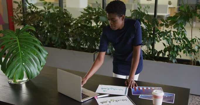 African american businesswoman standing using laptop going through paperwork in modern office