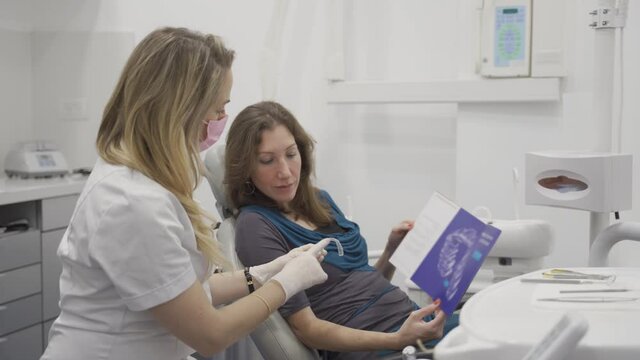 Orthodontist doctor holds invisible retainer showing patient a dental pamphlet or brochure in clinic pointing out details. Woman sits in clinic nodding and looking at a booklet for teeth alignment