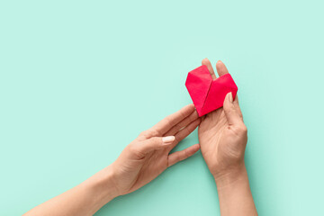 Hands with origami heart on color background