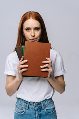 Upset sad young woman college student holding book and looking at camera on isolated gray background. Pretty redhead lady model wearing casual fashion clothes emotionally showing facial expressions.