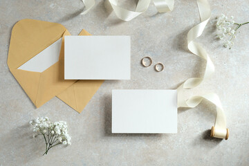 Wedding flat lay composition with blank paper card, envelopes, ribbon, flowers on stone table.