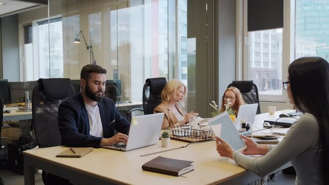 Medium Shot Of Group Of Friendly Multi-ethnic Coworkers Sitting At Common Desk Together, Working And Talking In Modern Open Space Office With Large Windows