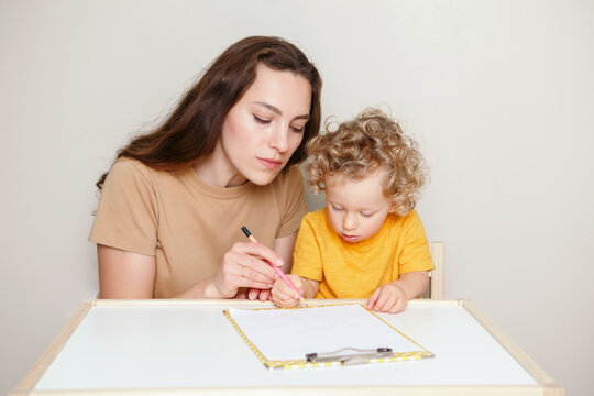 Mom drawing on paper together with baby boy toddler. Mother teaching kid to hold marker, pencil correctly. Early age kids education development learning. Family candid life at home.