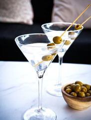 Well know cocktail at home, martini cocktail made with gin and vermouth, and garnished with olives poured in chilled martini cocktail glass on white table in the living room.