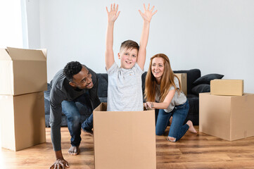 A multiracial family have fun in a new home, an African stepfather and caucasian mother driving school-age son in a cardboard box, they are laughing. Concept of moving in a new house