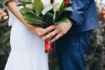 The bride in a white wedding dress and the groom in a blue plaid suit stand against each other and hold a large bouquet of white and pink roses with green foliage close up.