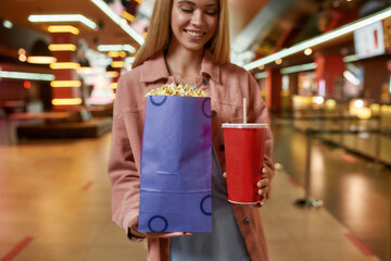 Cropped portrait of cheerful young woman holding big popcorn bag and soda while posing in front of cinema bar in a movie theater