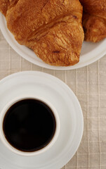 Flat lay composition of a cup of coffee on a white saucer and croissants on a beige tablecloth. French breakfast concept