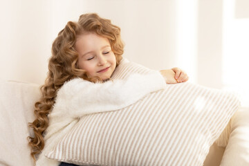 Naklejka premium happy little girl with long blond hair hugging a pillow and smiling, in beige interior at home. child is dreaming.