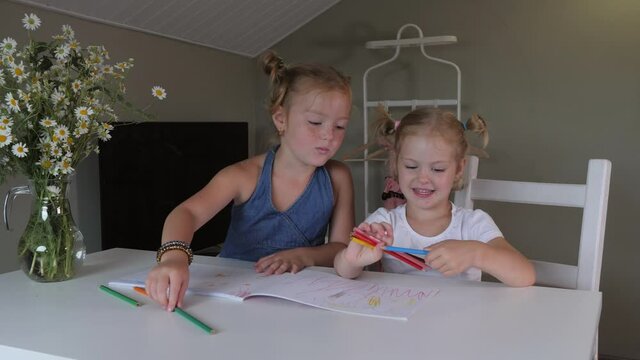 Two cute similar blonde girls with freckles are drawing with colored pencils in the album. Mischievous sisters are having fun at home. Carefree childhood.