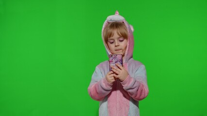 Excited little child girl in unicorn pajamas using, browzing on mobile cell phone, found out great big win news. Kid doing winner gesture isolated on chroma key background. Technology for children