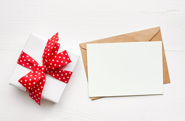 Greeting card mockup and gift box with red ribbon on white wooden background