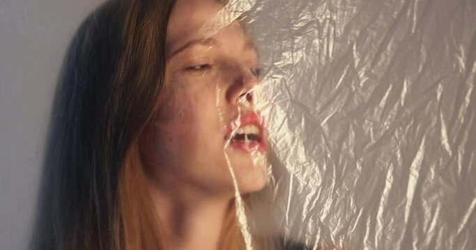 Shortness of breath. Defocused portrait. Respiratory disorder. Air pollution. Suffering woman trapped behind wrinkled transparent plastic polyethylene film isolated on gray background out of focus.