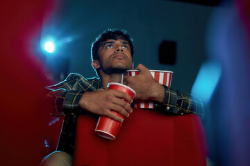 Low angle view of young guy holding a drink and popcorn basket while watching movie alone in empty theater auditorium