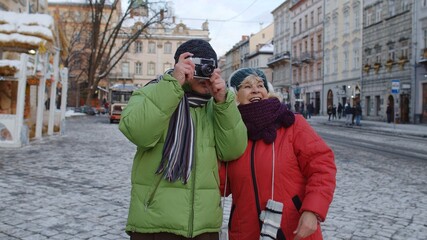 Fototapeta na wymiar Stylish senior elderly couple tourists grandmother and grandfather traveling, taking photo pictures on retro camera on winter city street. Lovely mature pensioners family vacations Christmas holidays