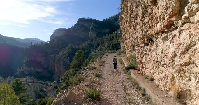 dron vuela hacia atr&aacute;s adolescente con bast&oacute;n camina en senda junto a barranco monta&ntilde;oso