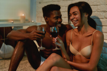 Young happy african couple, half naked man and woman drinking wine, talking and laughing while sitting on the bed at home
