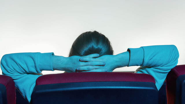 Back view of relaxed guy leaning on the back of chair and keeping his hands over the head while watching a movie alone in empty theater auditorium