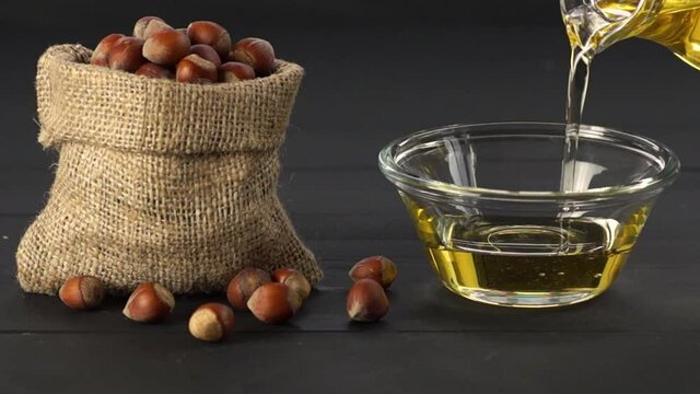 Hazelnuts, filbert in burlap sack and hazelnut oil in glass bowl pouring from bottle on black wooden table