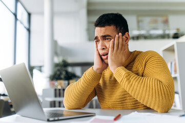 Confused hispanic male freelancer, student or coworker surprised looks at laptop while working or studying online, he got bad news or an unexpected message