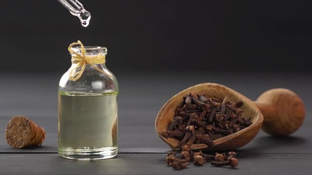 Glass bottle of essential clove oil and cloves in wooden shovel or spoon on black wooden table, oil dripping