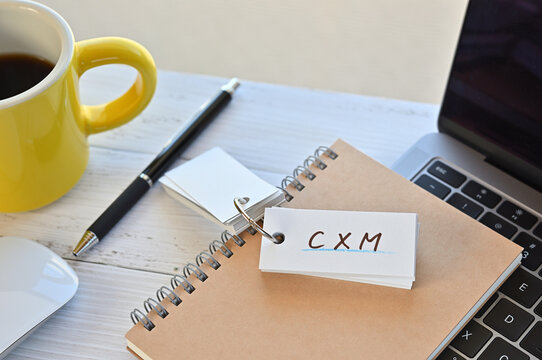 There's a word book on a desk with a cup of coffee and laptop pc. The word CXM is written in it. It's an abbreviation for Customer Experience Management.