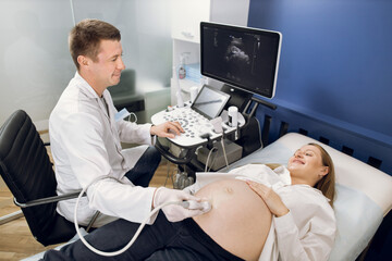 Smiling male doctor obstetrician, examining belly of happy pregnant woman by ultrasonic scan. Cheerful man doctor doing ultrasound procedure for his patient, looking each other. Top angle view