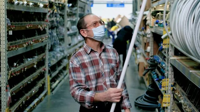 An elderly man wearing a face mask and a plaid shirt picks out a pipe in the plumbing section of a construction store. Portrait. Close-up. The camera moves around. Epidemic, coronavirus