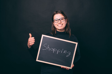 Young girl wearing glasses is holding a chalkboard with the word shopping, while showing thumb up over black background.