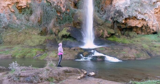 Mujer con ropa deportiva rosa junto a catarata vista a&eacute;rea de dron