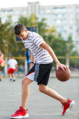 Young cute boy plays basketball at the outdoor streetball court on a sunny summer day. Teenager player in action dribbling the ball. Hobby, active lifestyle, sports for kids.