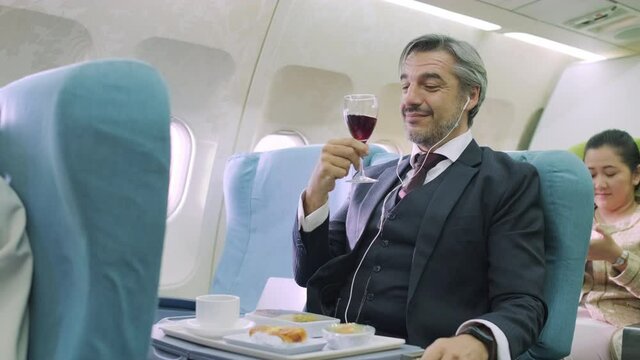 Senior businessman or passenger listen music with earphone, sitting in the first class of airplane. Enjoying the flight and drinking red wine, feeling smile and happy. Stewardess serve food and drink