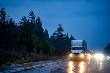 Big rig white semi truck with turned on head lights transporting cargo in semi trailer running on...