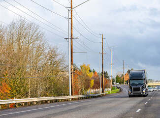 Black big rig semi truck with semi trailer running on the local road with power line poles on the...