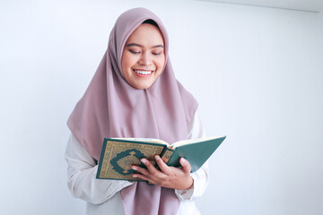 Young Asian Islam woman wearing headscarf is praying or read Quran - the holy book of Islam with smile and serious face. Indonesian woman on gray background