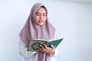 Young Asian Islam woman wearing headscarf is praying or read Quran - the holy book of Islam with smile and serious face. Indonesian woman on gray background