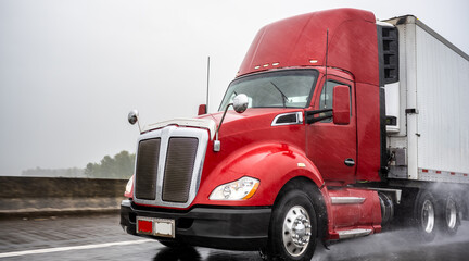 Day cab big rig red semi truck with roof spoiler transporting cargo in refrigerated semi trailer running on the wet rain highway at raining weather
