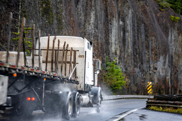 Industrial big rig classic white semi truck with flat bed semi trailer running on the wet winding mountain road with rock cliff  at raining weather