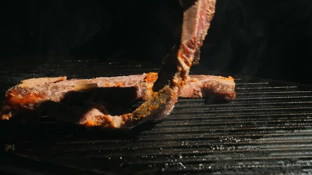 This slow motion video shows fresh short rib steaks being placed on a cast iron grill to cook with metal tongs.