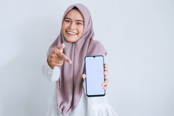 Young Asian Islam woman wearing headscarf is pointing finger on white screen of phone with smile and happy feeling. Indonesian woman on gray background