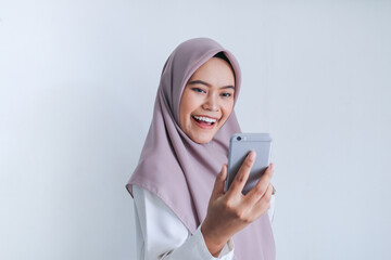 Young Asian Islam woman wearing headscarf is happy and excited celebrating in what she see on the smartphone. Indonesian woman on gray background